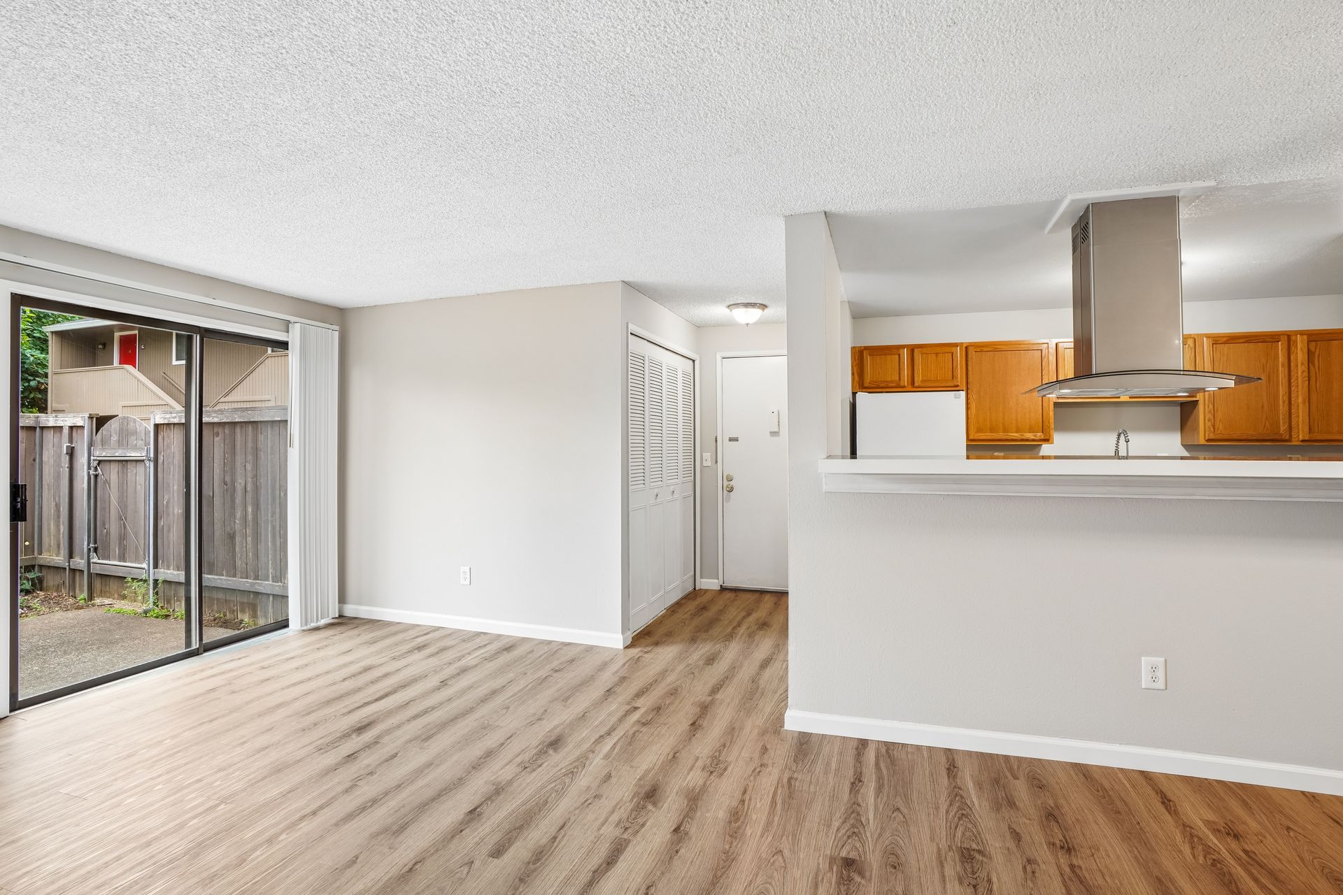 Empty apartment living room with wood floors, sliding glass door, and open kitchen with maple cabinets