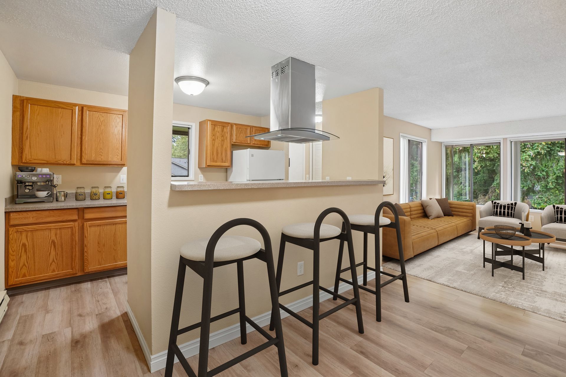 Open-concept living room and kitchen with bar stools, tan sofa, and large windows.