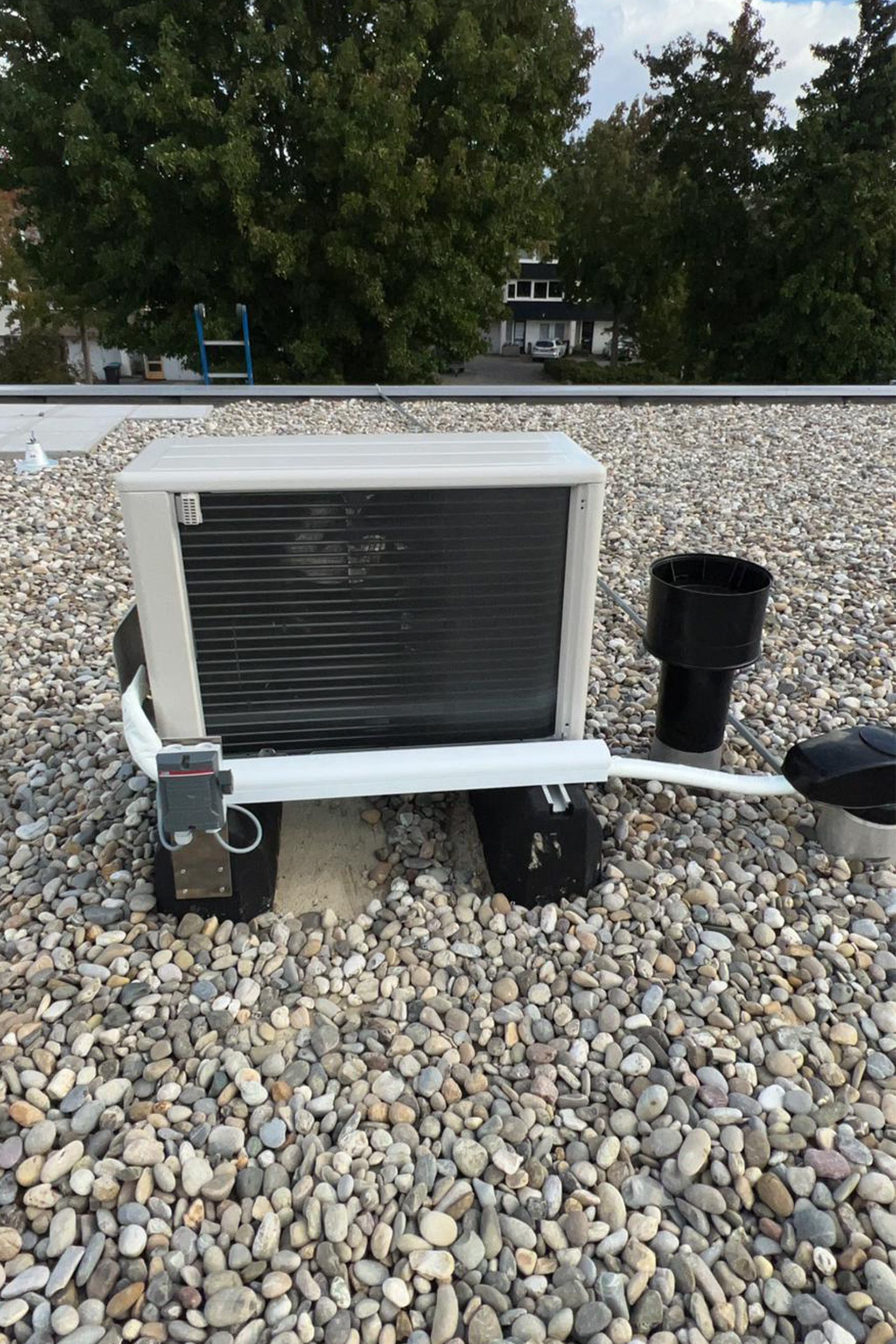 Airconditioningunit op een grinddak, met aangesloten leidingen, naast een zwarte afvoerbuis.