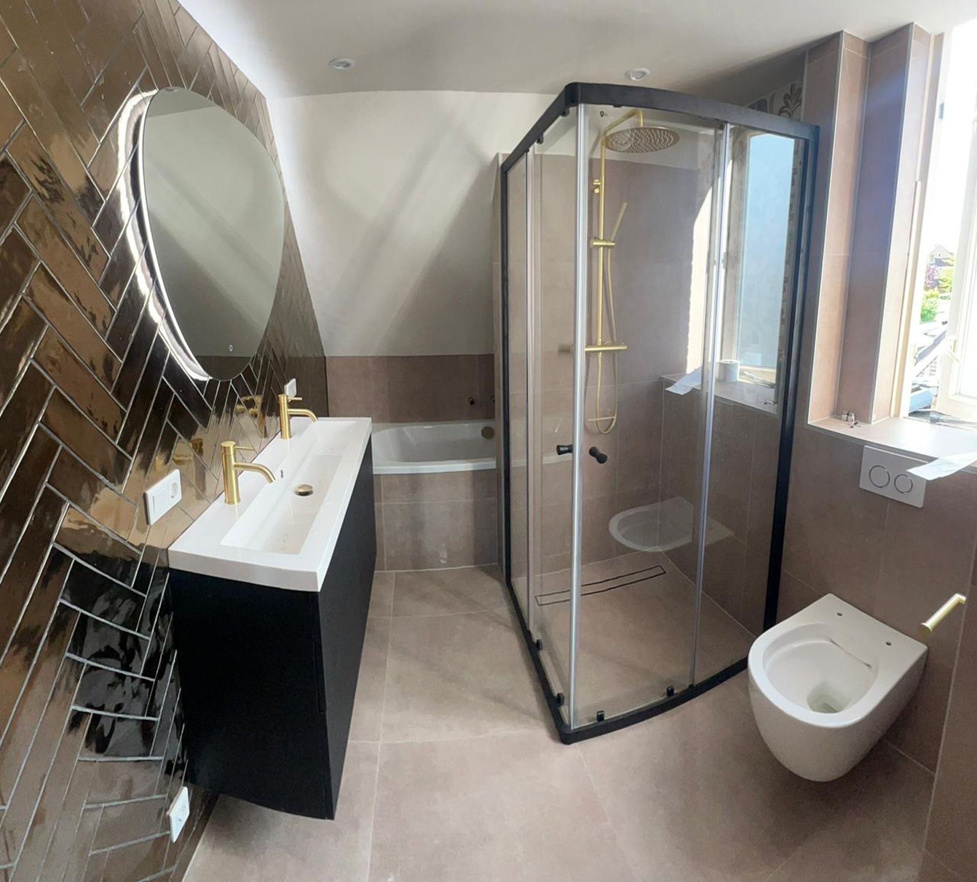 Moderne badkamer met zwart badmeubel, gouden kranen, glazen douchecabine, visgraattegels en een wandtoilet.