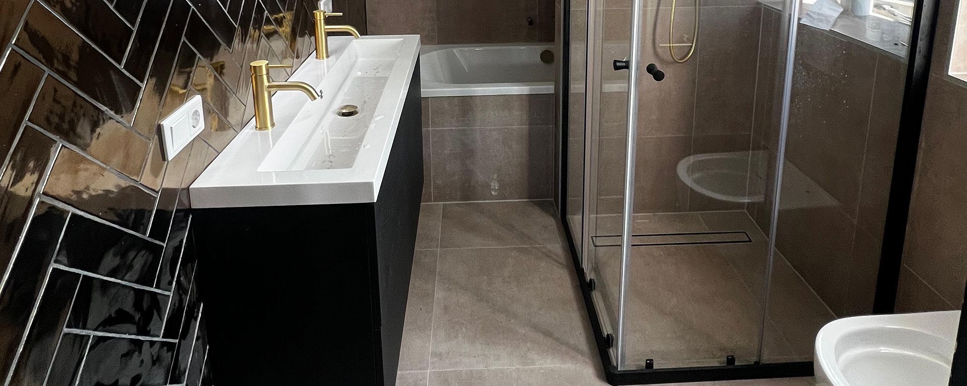 Badkamer met zwart badmeubel, gouden kranen, visgraattegels, douche en bad.