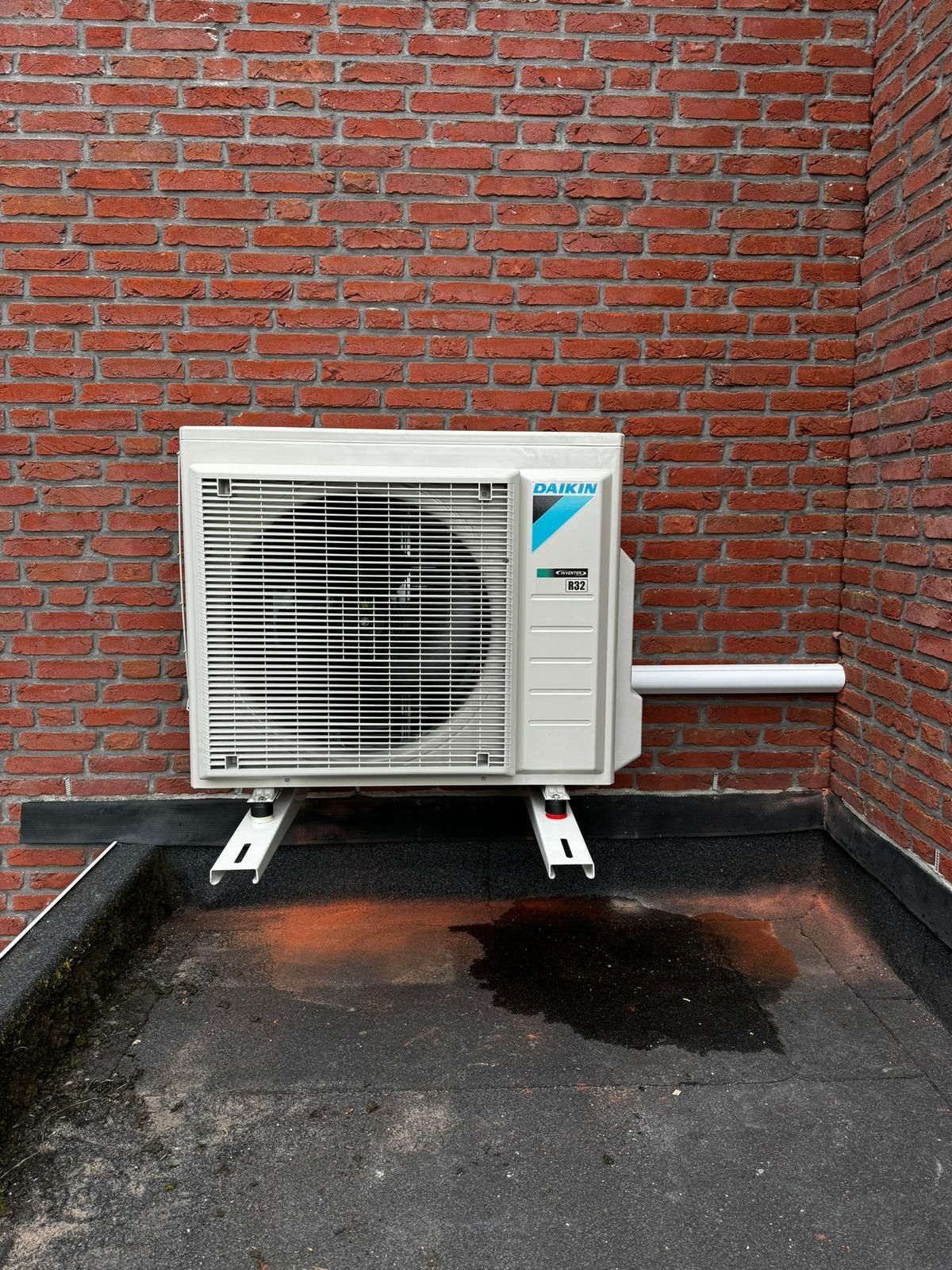 Een airconditioner is gemonteerd op een plat, zwart dak tegen een bakstenen muur.