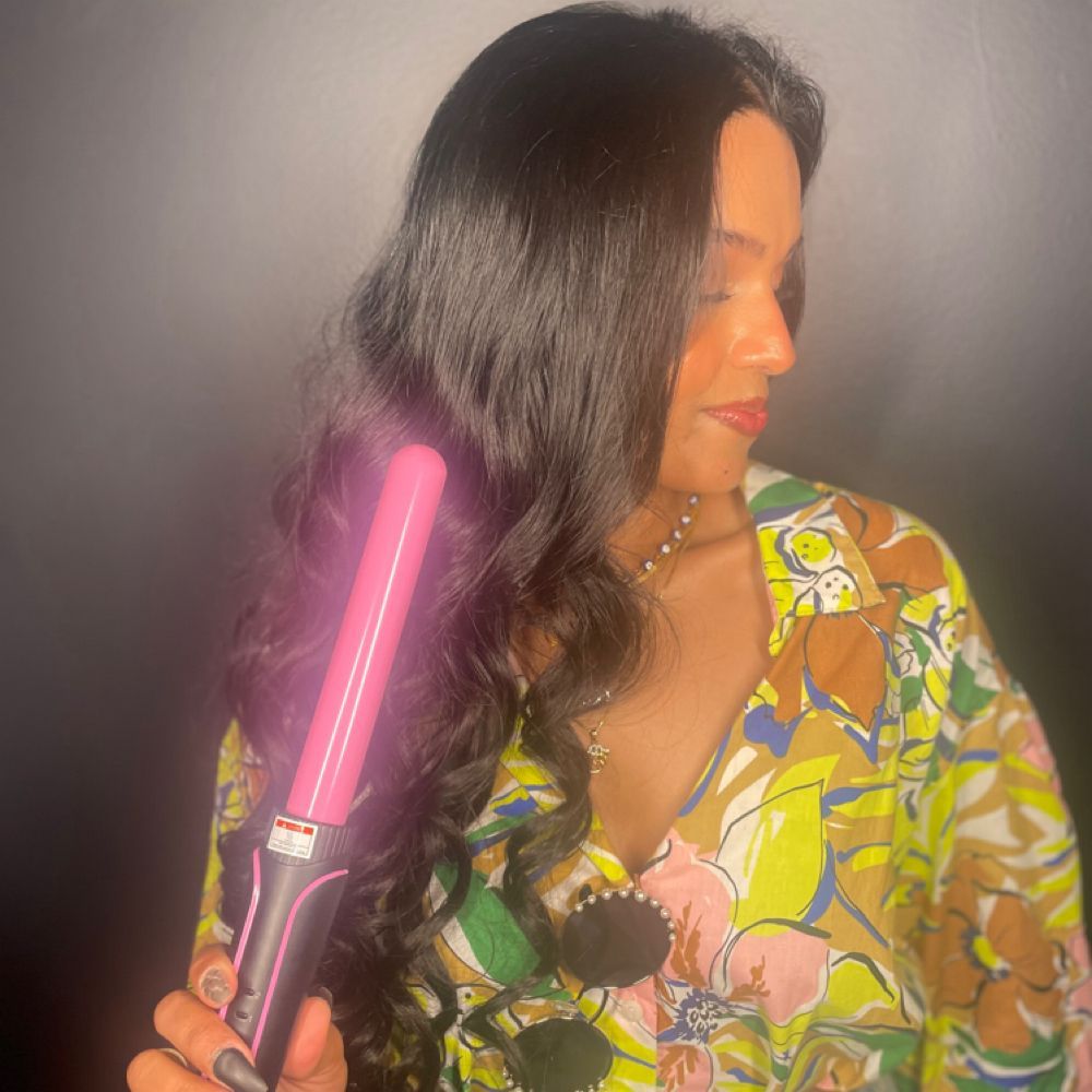 Woman holding a pink glowing hair straightener beside her long dark hair against a gray background