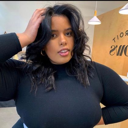 Person in a black top posing indoors with one hand in their hair in a bright café setting