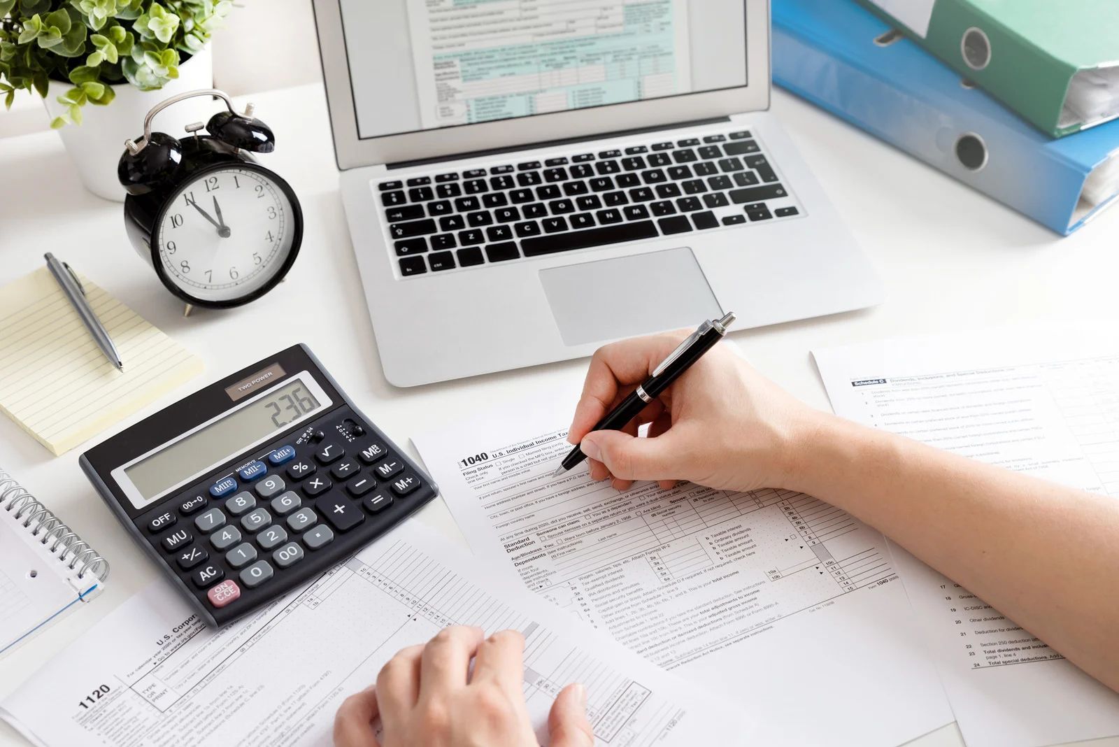 Hand writing on financial paperwork beside a calculator, laptop, alarm clock, and binder folders