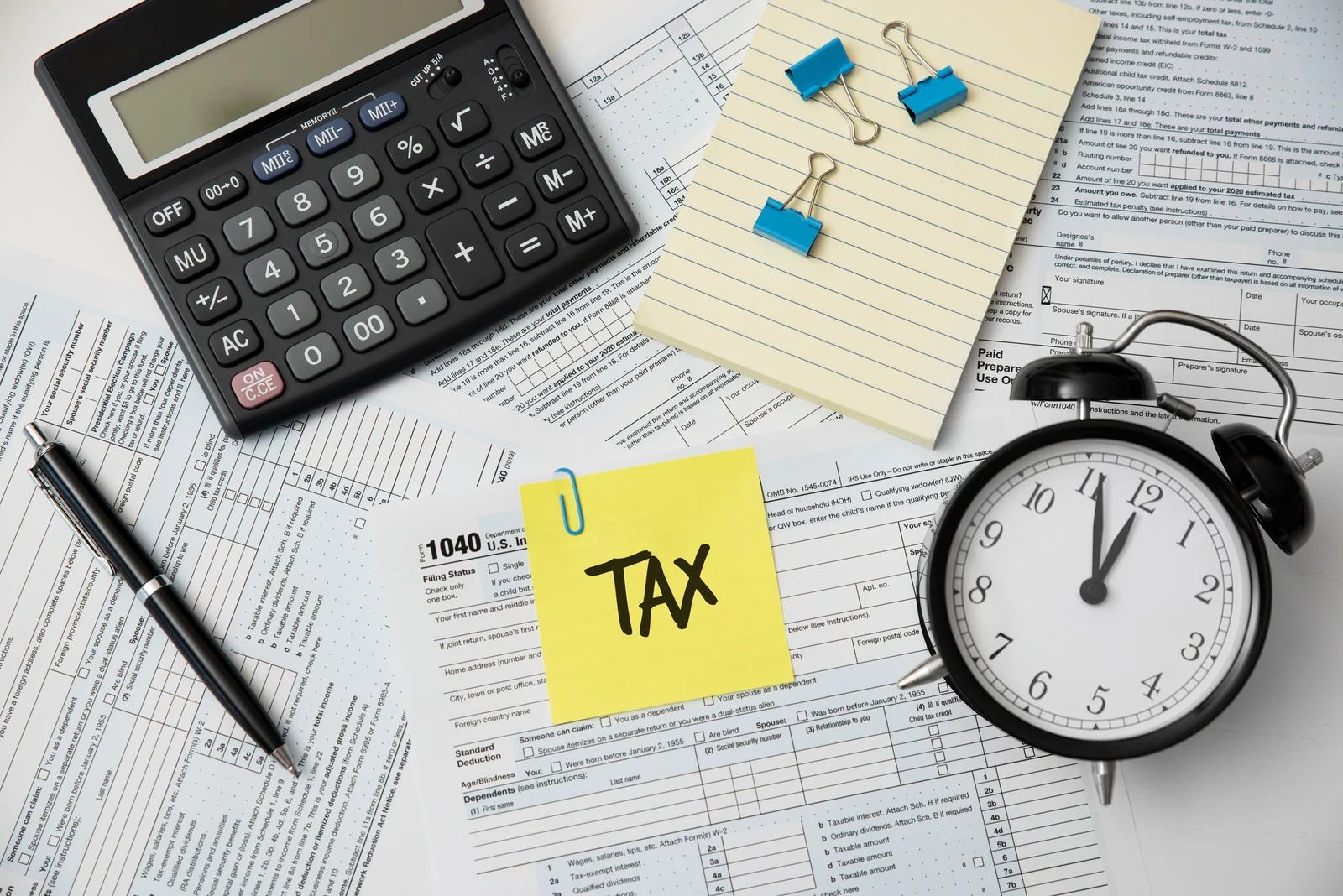 A calculator, a black alarm clock, and a pen on tax forms, with a yellow sticky note labeled