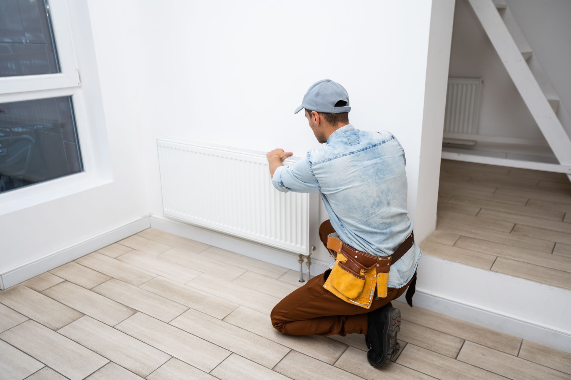 A technician kneeling beside a wall radiator, tool belt on, doing skilled heating repair work. A technician kneeling beside a wall radiator, tool belt on, doing skilled heating repair work.