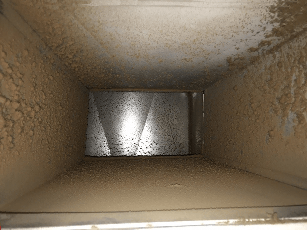 Inside of a dusty HVAC duct; light reflects off a central vent, obscuring the far end.
