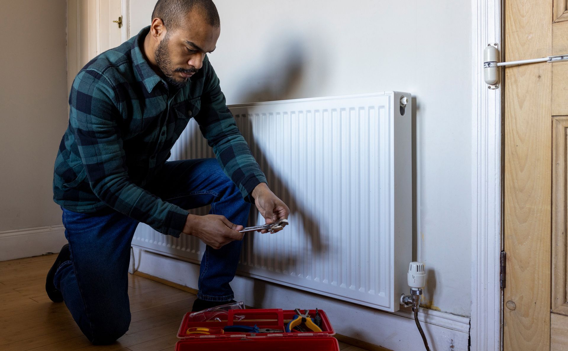 A person kneeling by a white radiator, using a tool from a toolbox.