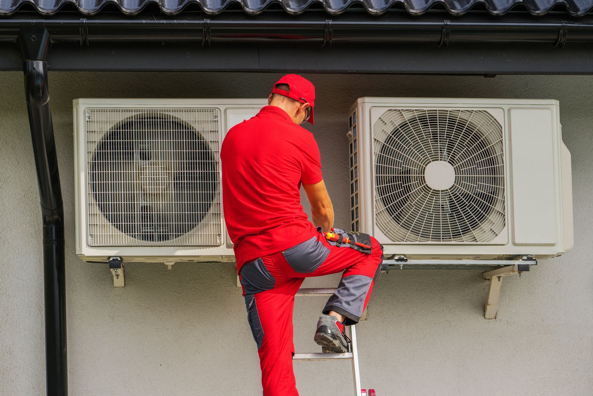 Male technician in red uniform performs residential heating repair on exterior HVAC unit. HVAC technician in red overalls on a ladder, working on two outdoor air conditioning units.