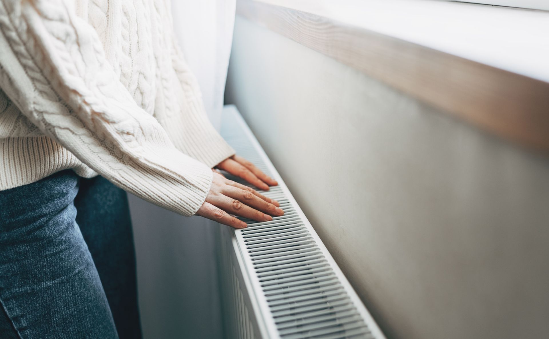 Woman checking heating radiator in cold apartment. Person in a cable-knit sweater and jeans touching a white radiator near a window.