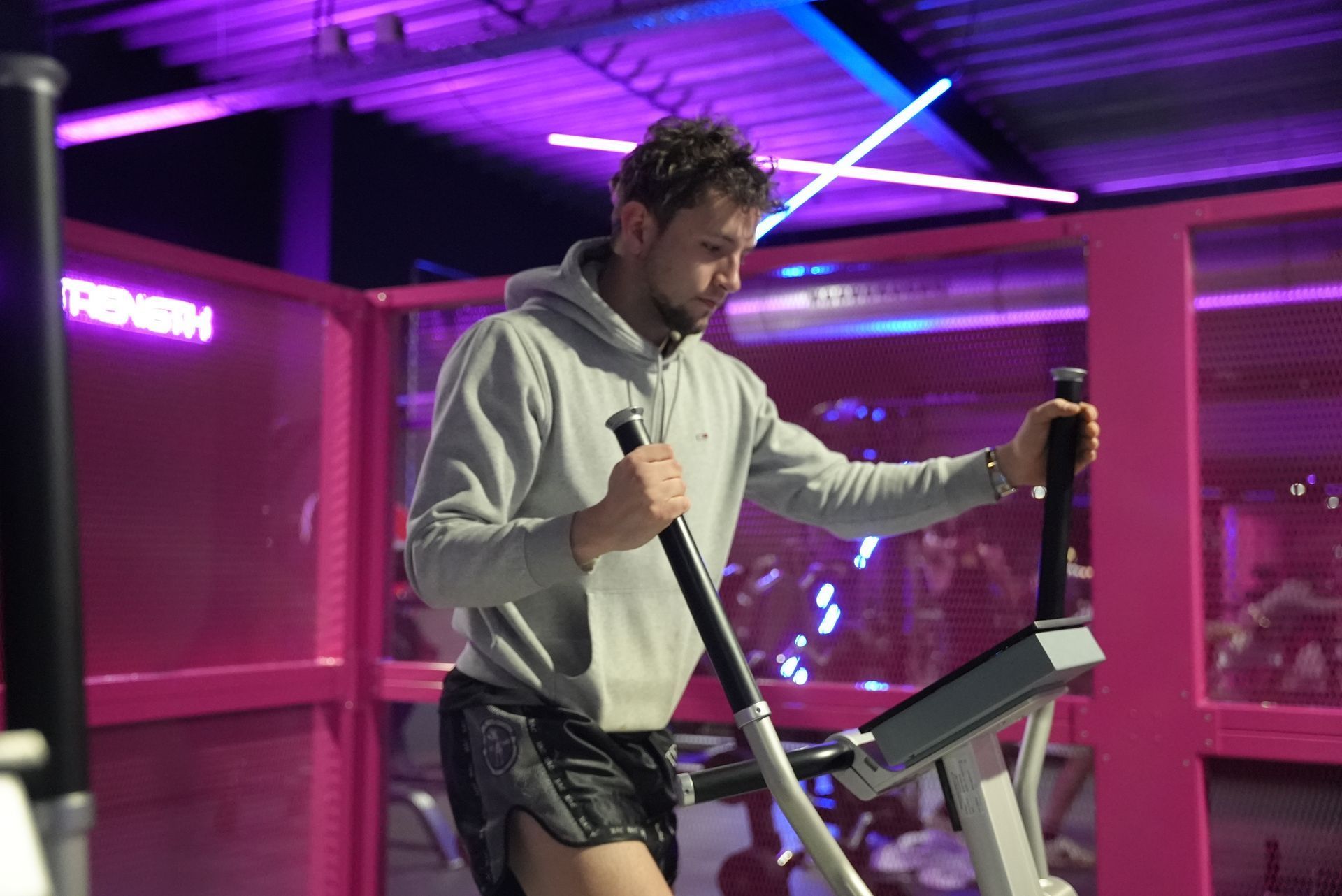 Man in grey hoodie using an elliptical machine in a gym with pink and purple lighting.