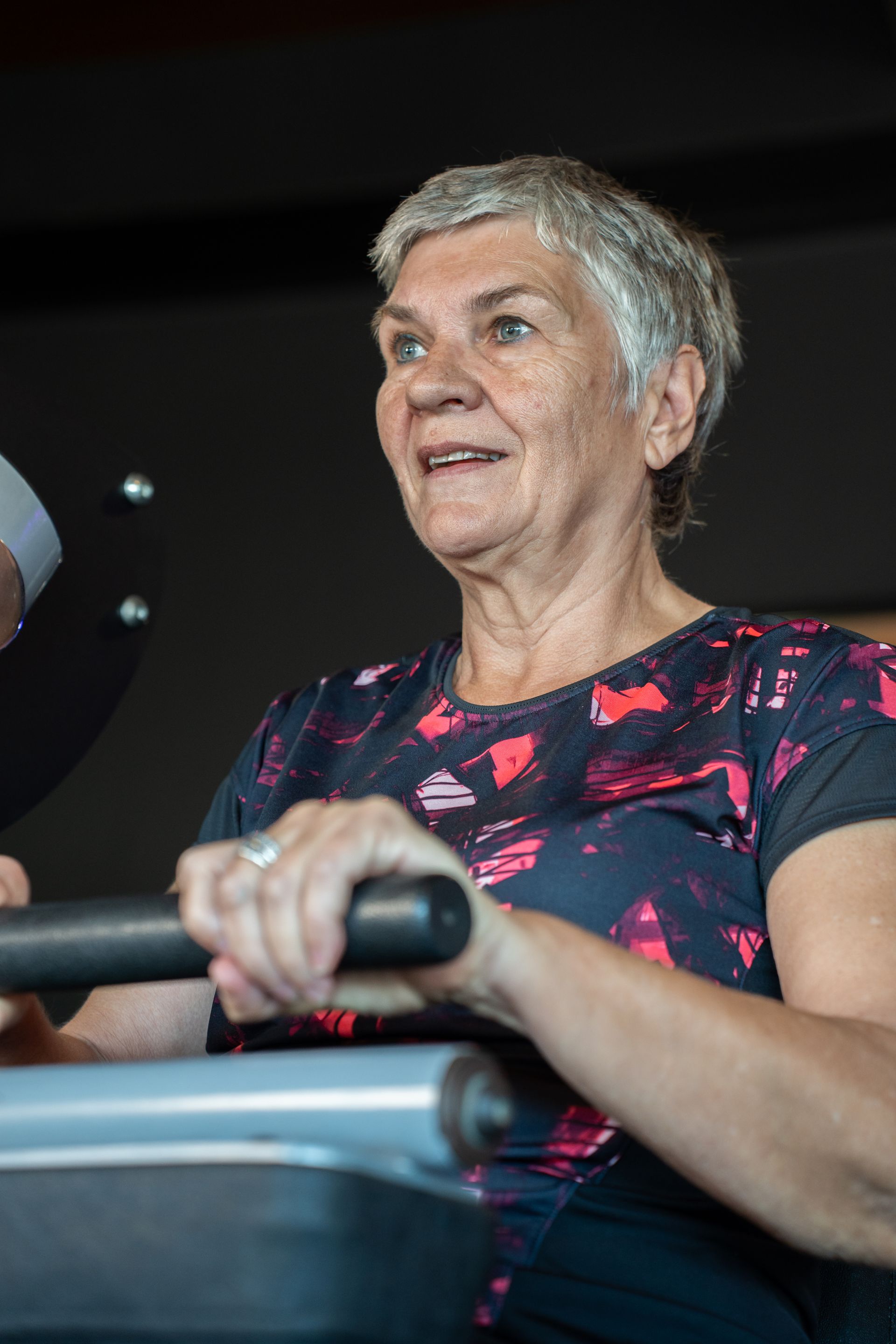 Vrouw die aan het sporten is op een apparaat, gekleed in een donker shirt met patroon en omhoog kijkend.