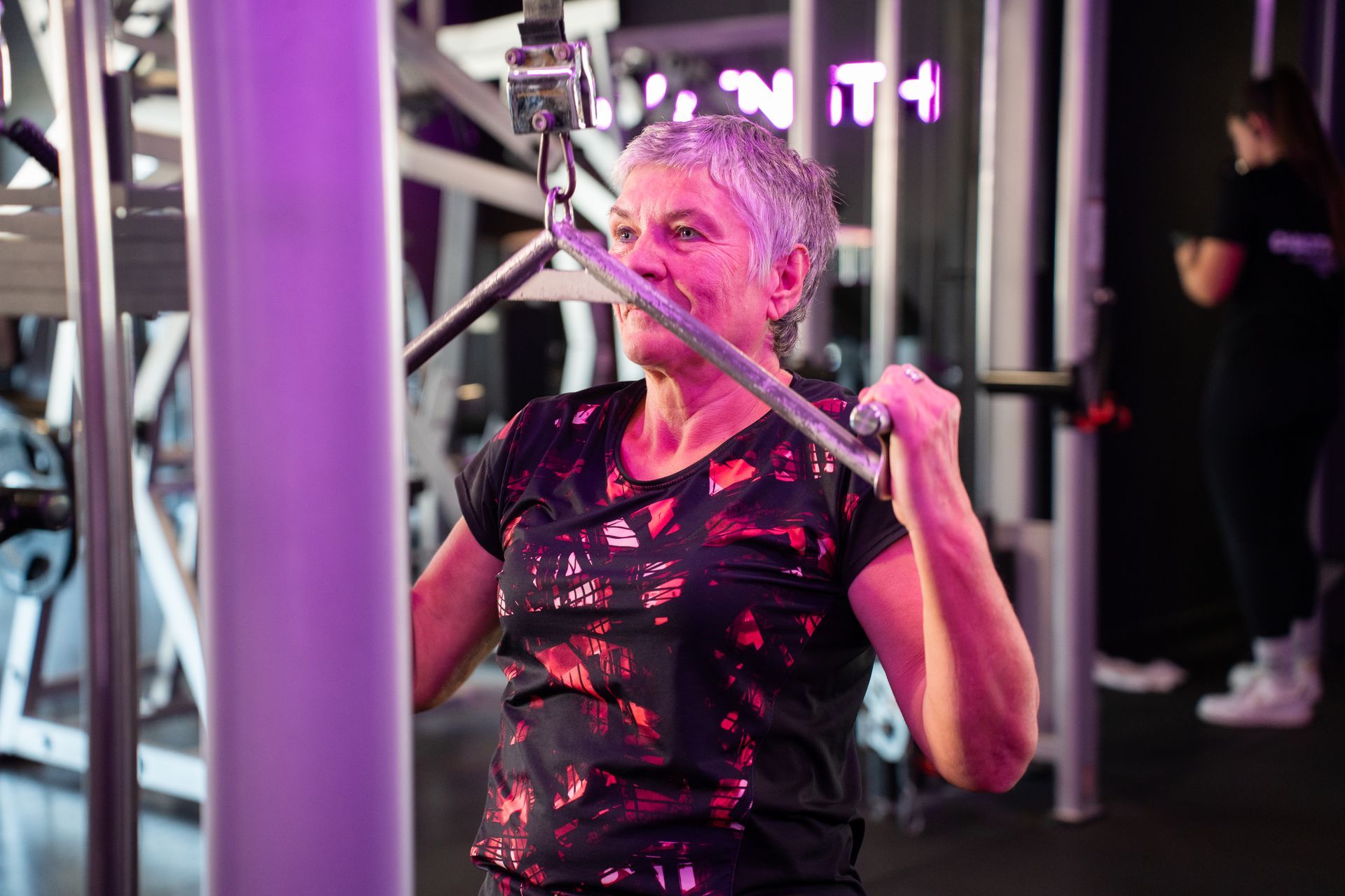 Vrouw die een lat pulldown-apparaat gebruikt in de sportschool, lachend onder roze verlichting.