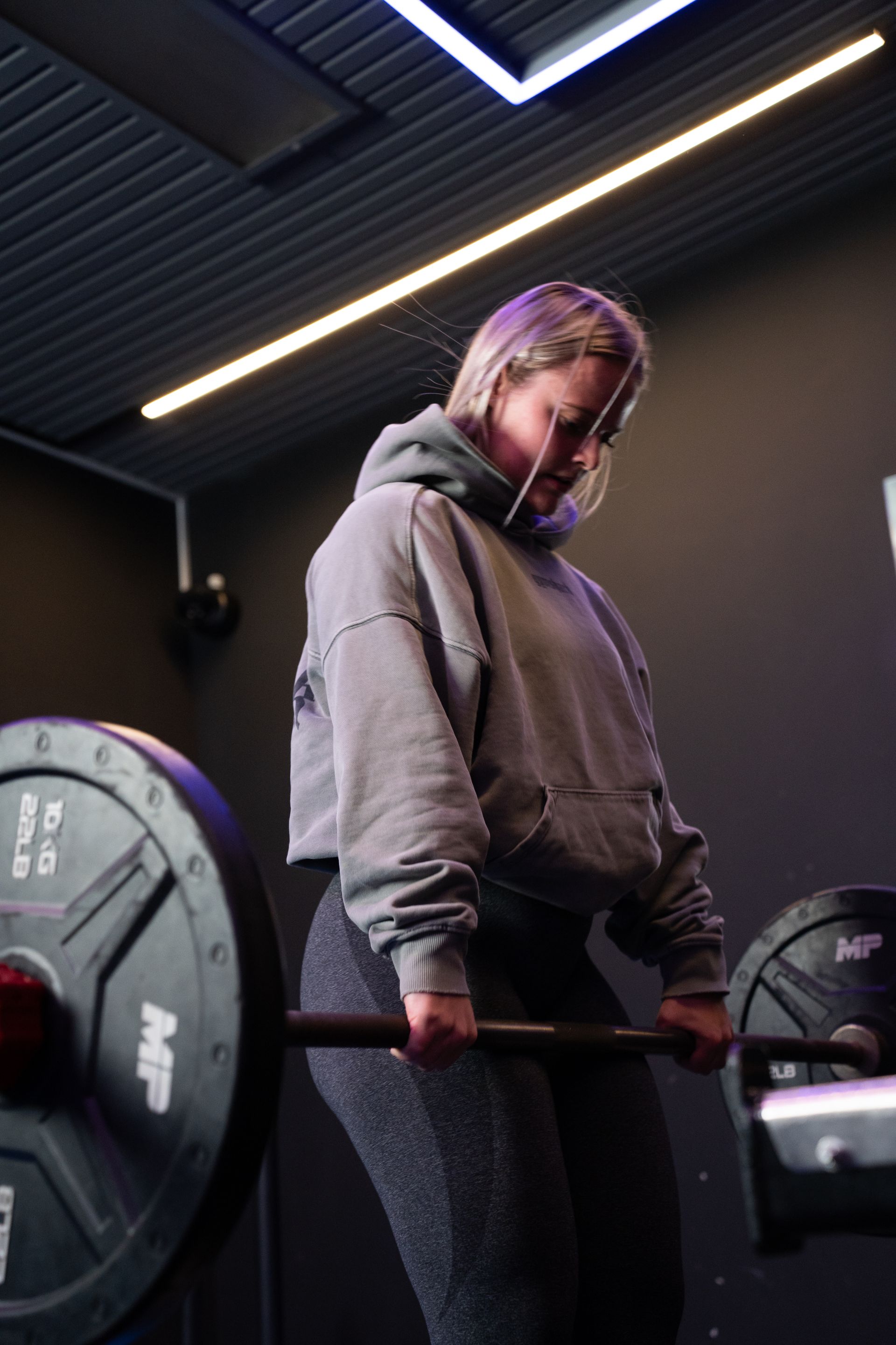 Een persoon die een halter optilt in een sportschool, gekleed in een grijze hoodie en een zwarte legging.