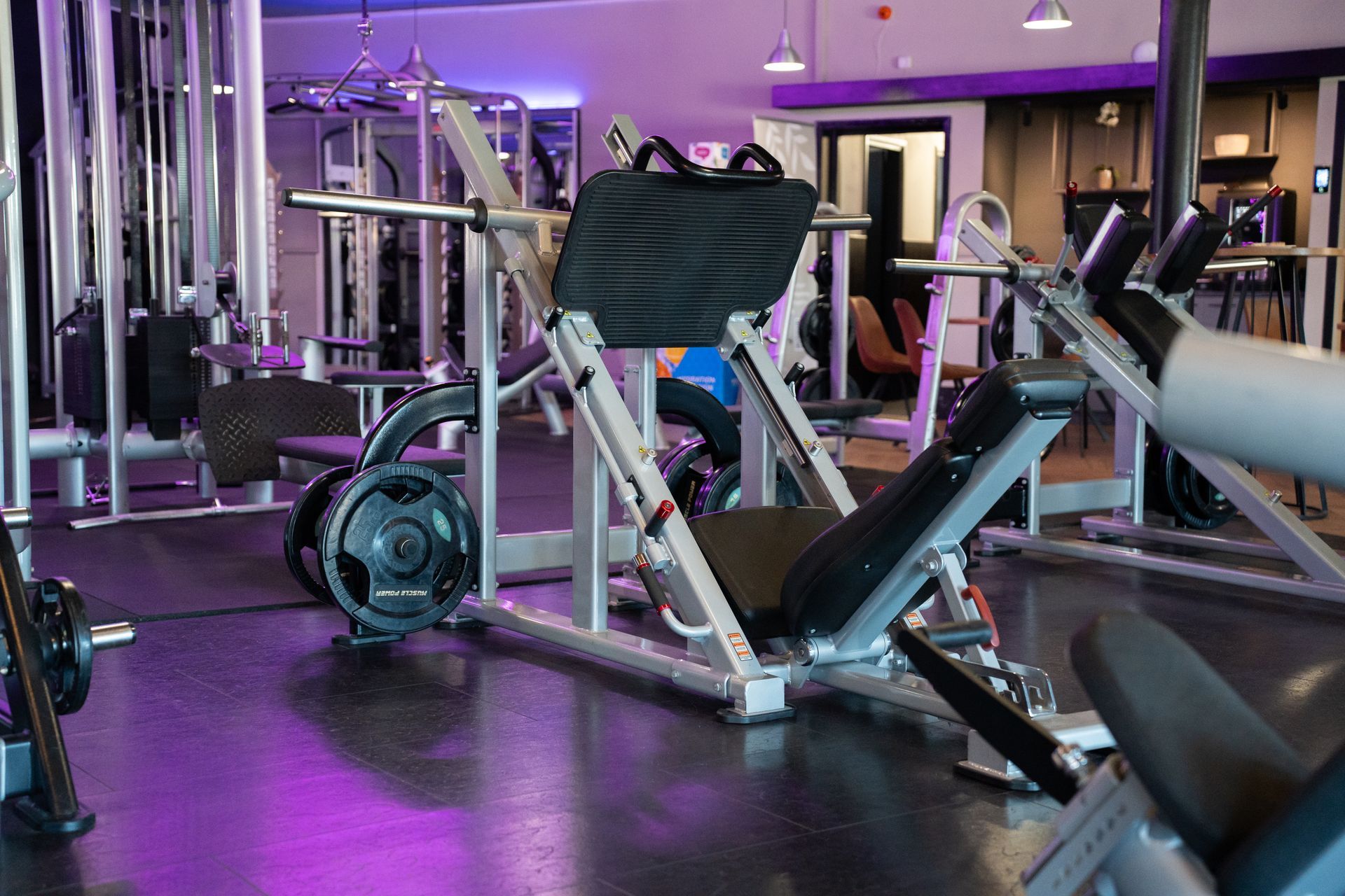 Paars verlichte sportschool met gewichtsapparaten, banken en cardio-apparatuur op een donkere vloer.