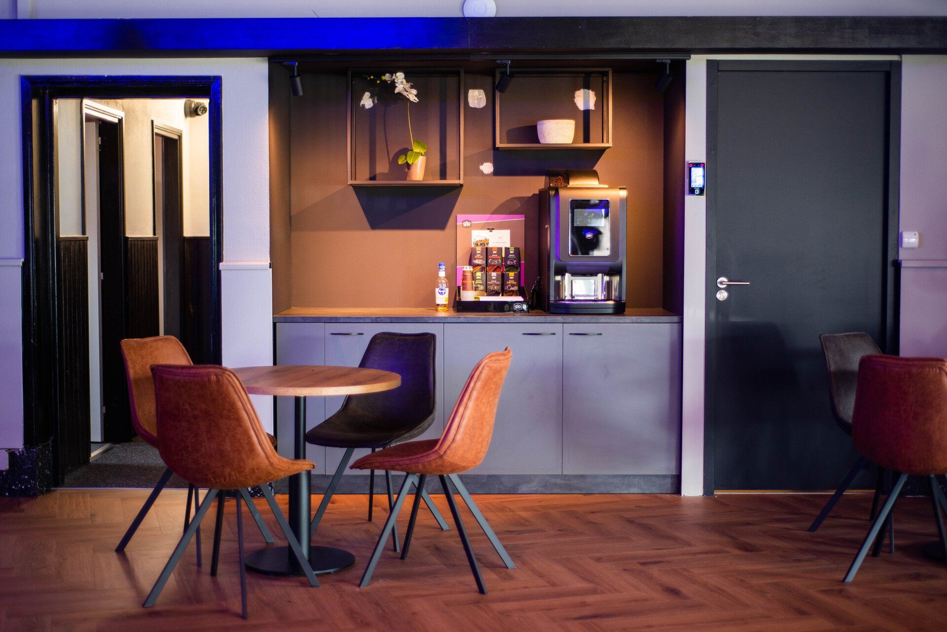 Moderne lounge met ronde tafel, oranje stoelen, koffiezetapparaat en donkere deuren onder paarse verlichting.