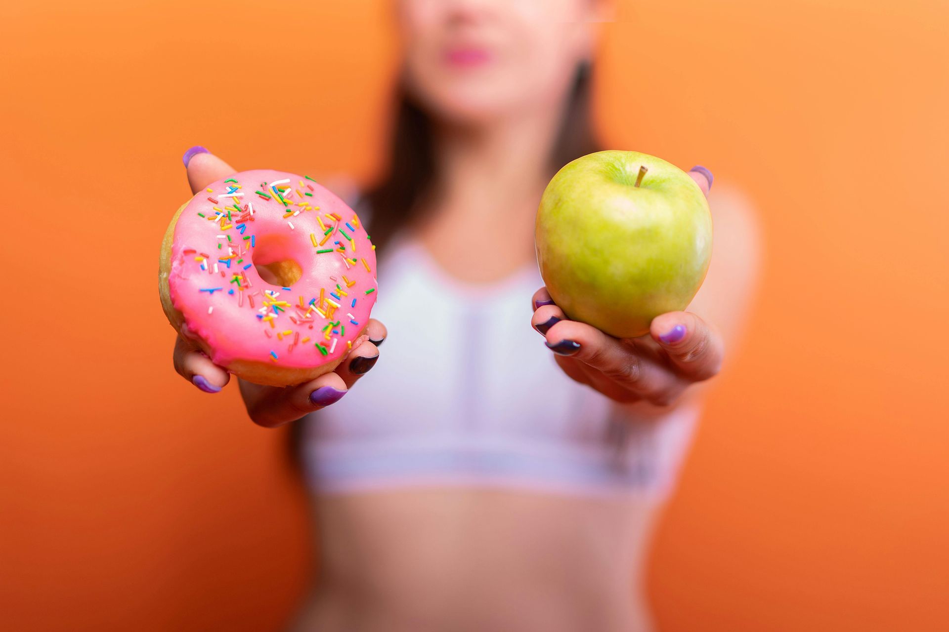 Woman holding a pink donut and a green apple, making a food choice. Orange background.