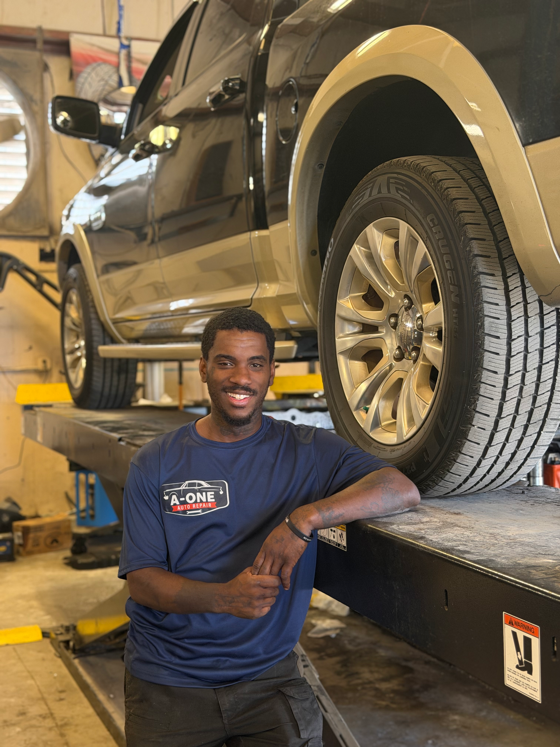 Smiling person leaning on a car in a garage, wearing a dark hoodie and jeans.
