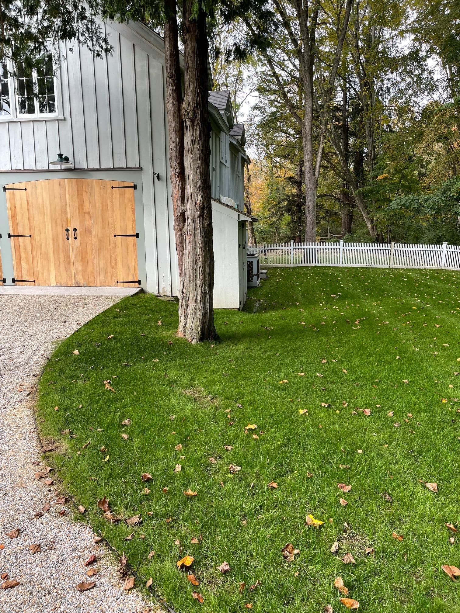 Gravel driveway beside a gray building with a wood door, green lawn, trees, and a white fence.