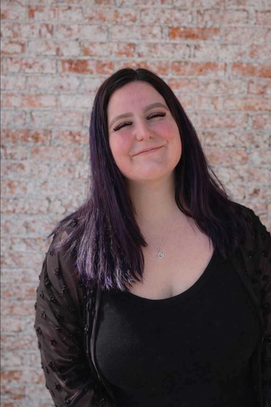 Smiling person with long dark hair standing against a brick wall, wearing a black top and sheer cover-up