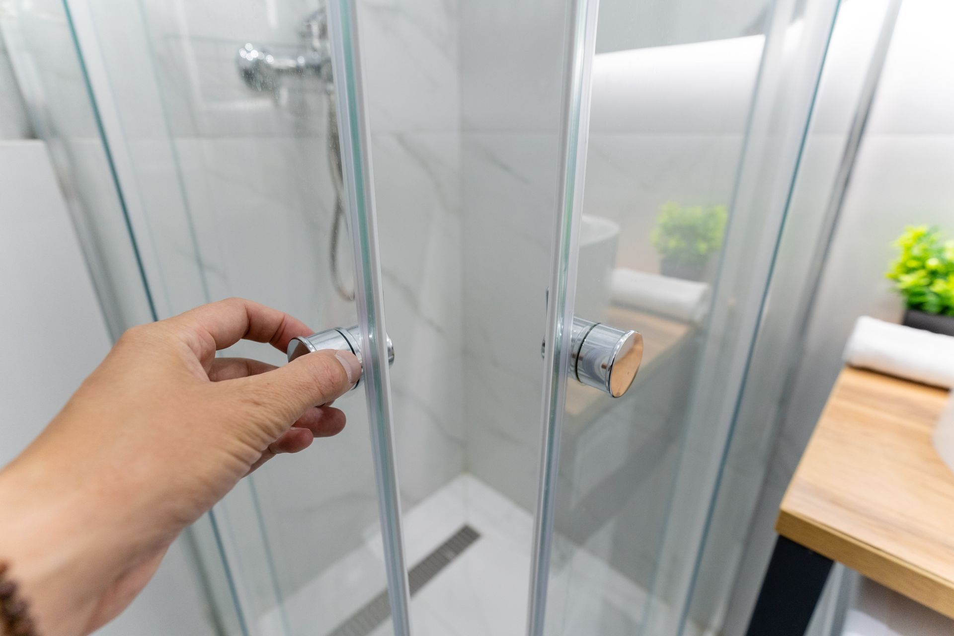 Hand opening a glass shower door with chrome handle in a bright bathroom