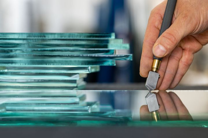 Hand scoring a glass panel with a cutter beside stacked glass sheets