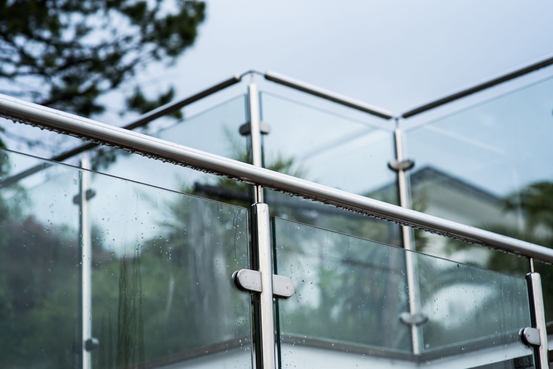 Glass balcony railing with metal posts and blurred greenery in the background