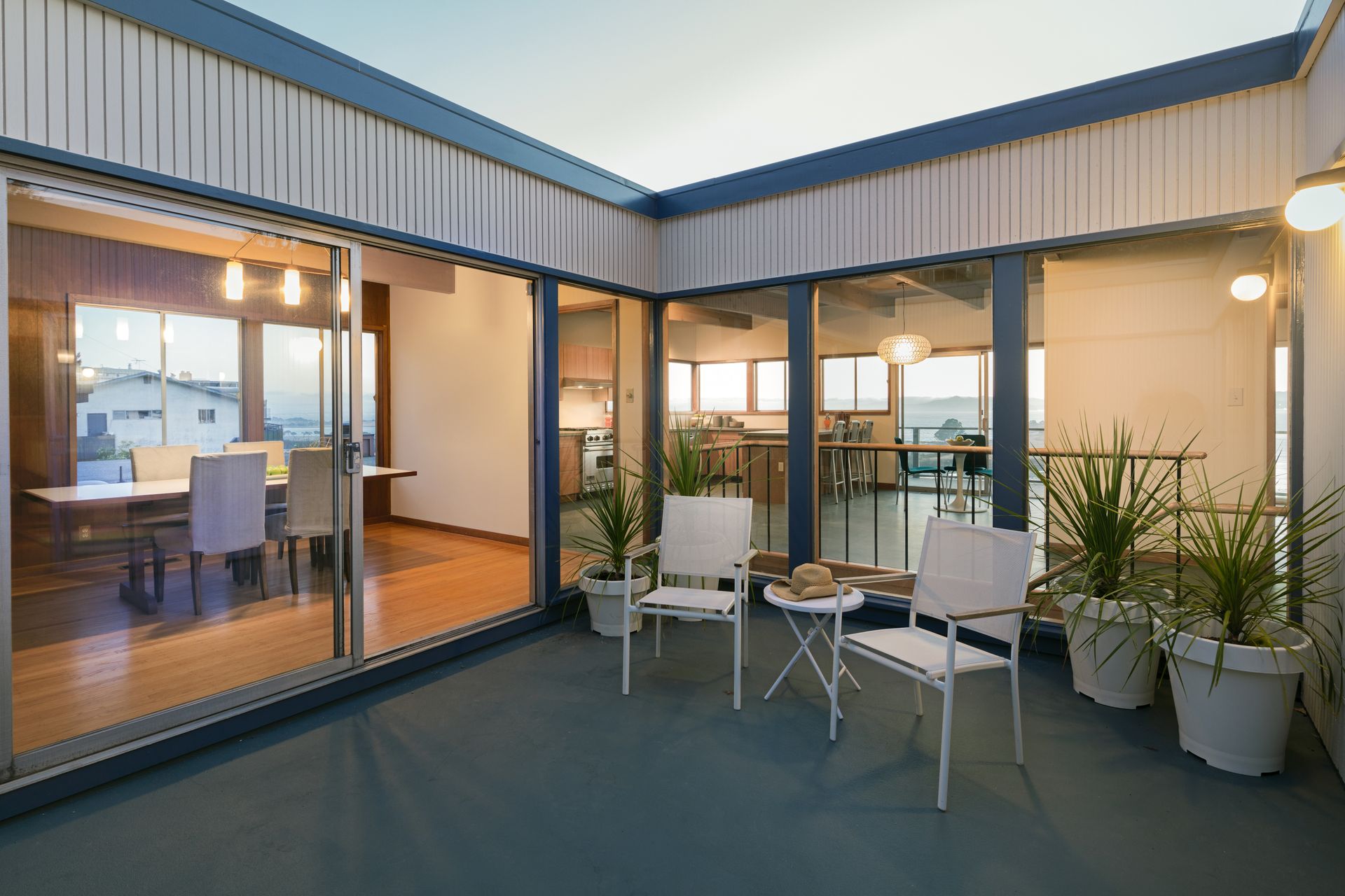 Patio seating area beside a glass-walled room, with white chairs, potted plants, and warm interior lights.