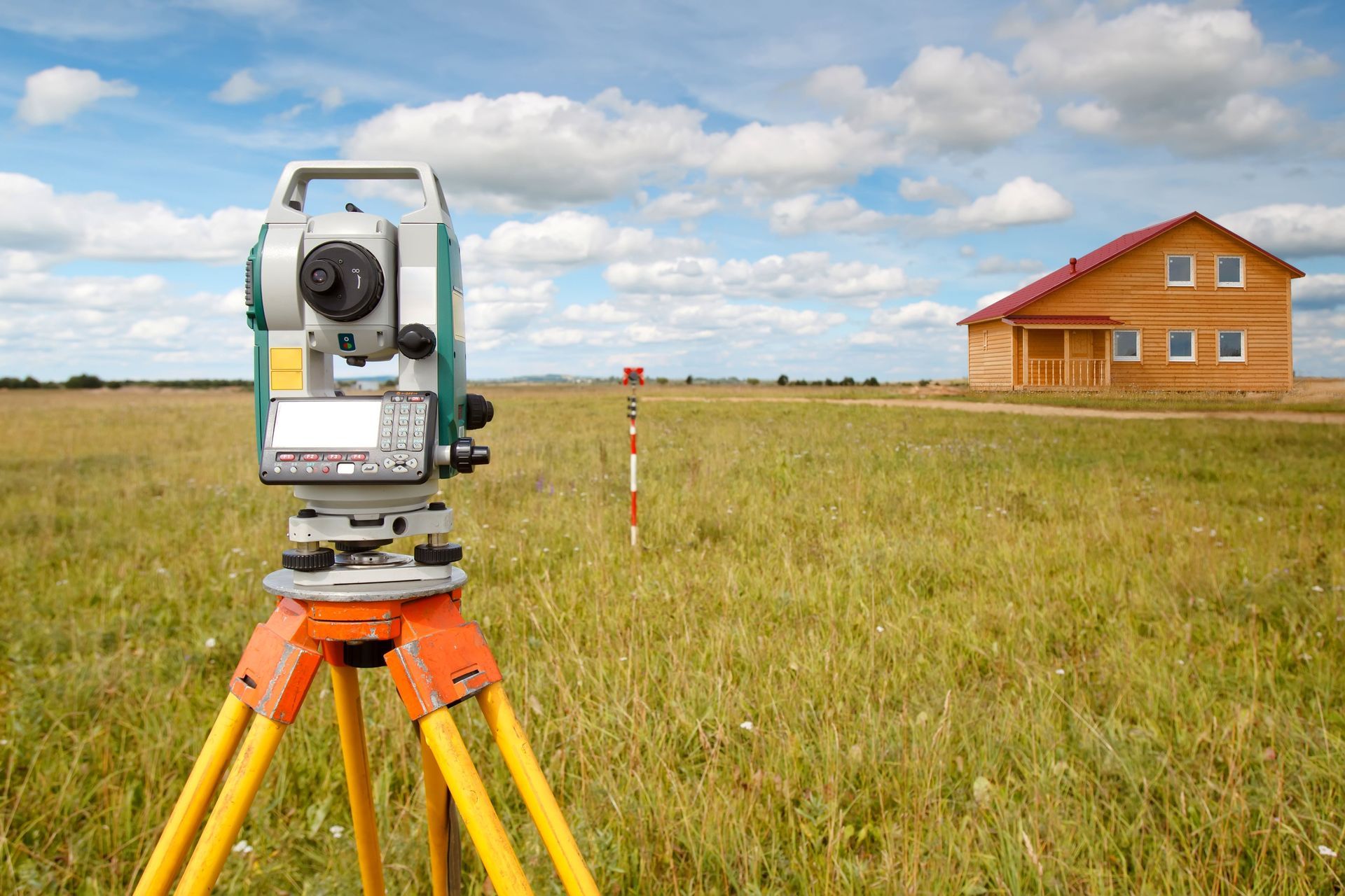 Surveying instrument on tripod in grassy field with wooden house in the distance under a cloudy sky