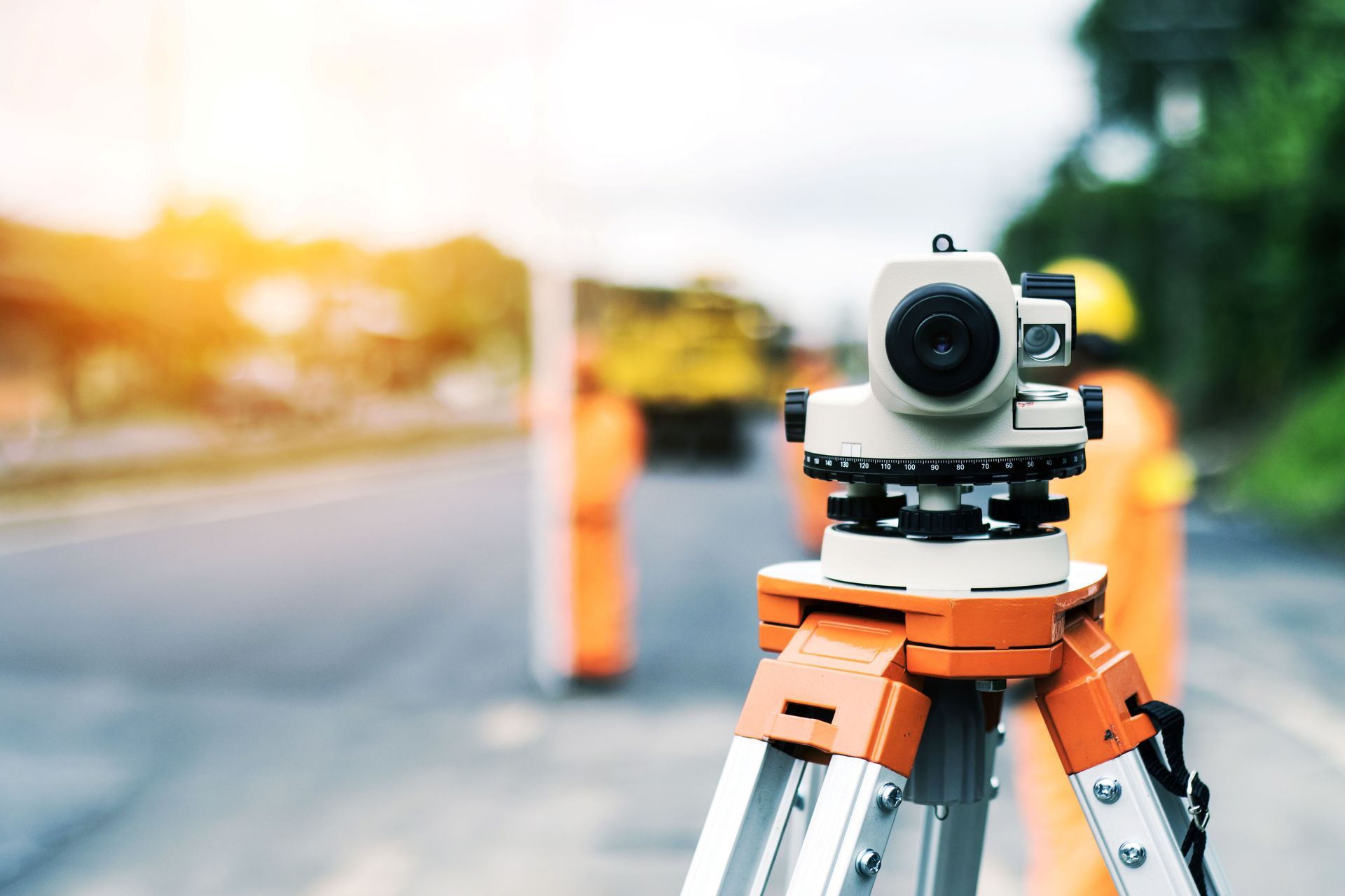 Surveying camera on tripod beside a sunlit road, with blurred barriers and warm evening light.