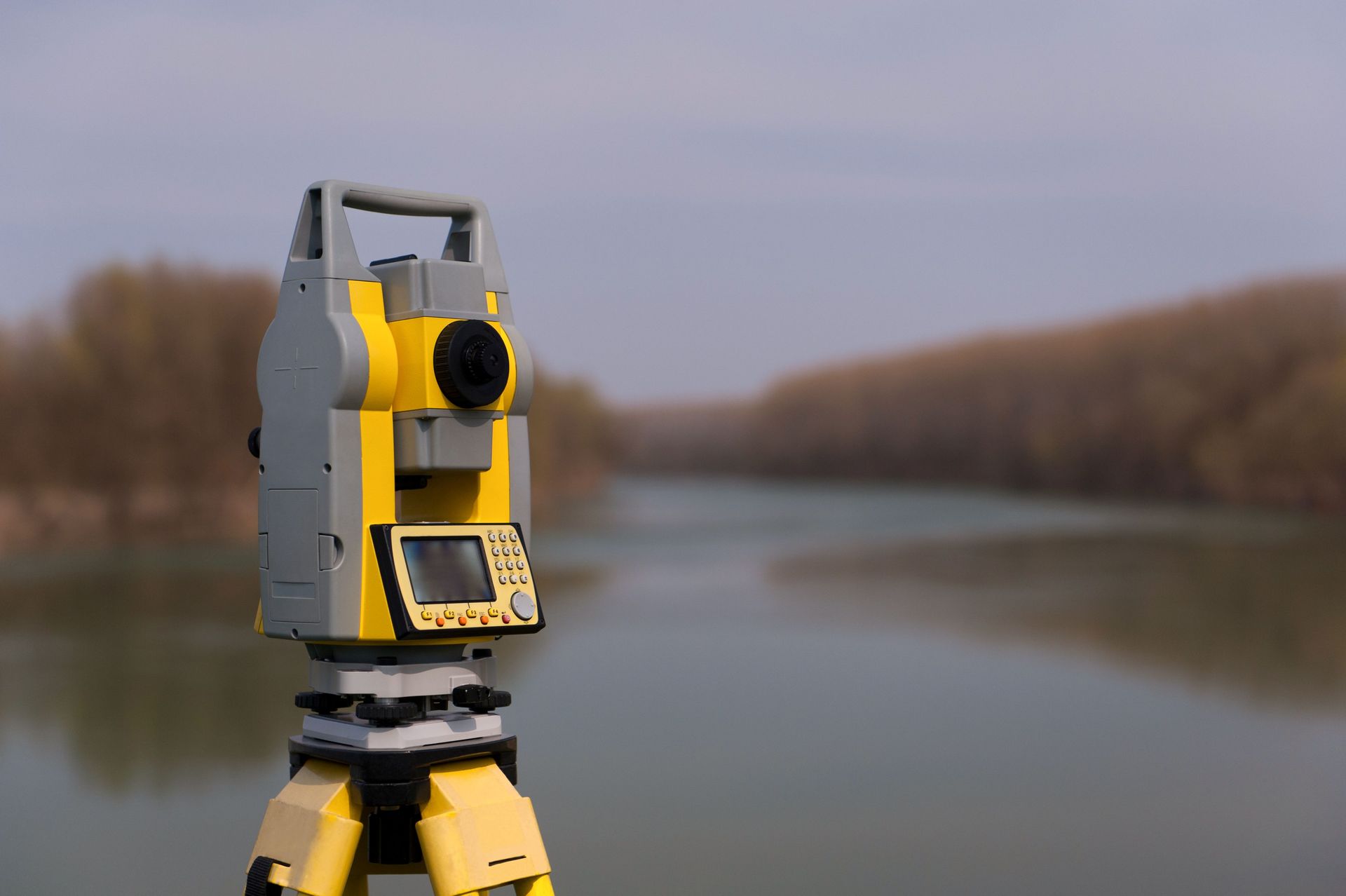 Yellow surveying instrument on a tripod beside a calm river under an overcast sky