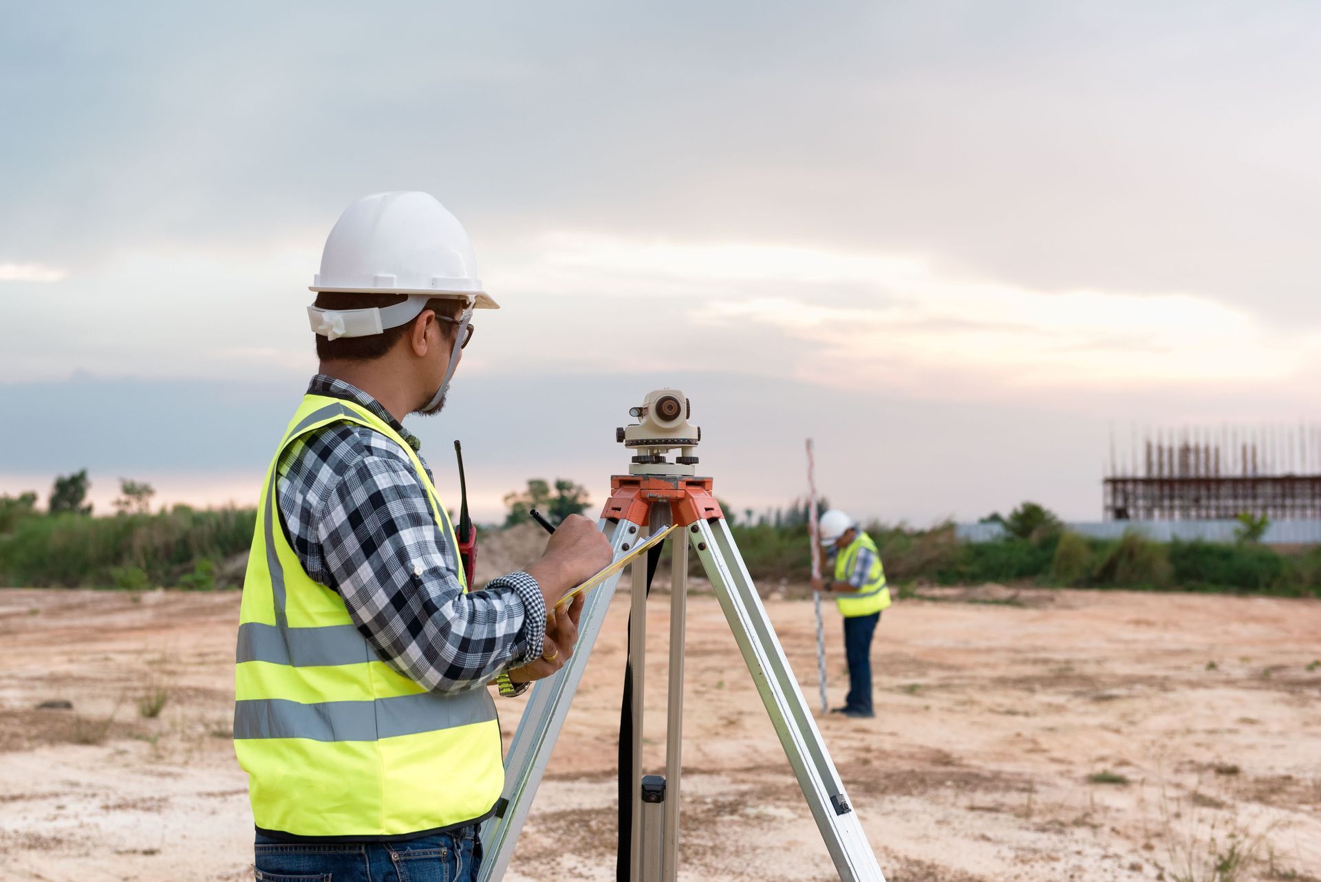 Surveyor in safety vest using tripod equipment on a sandy construction site at dusk
