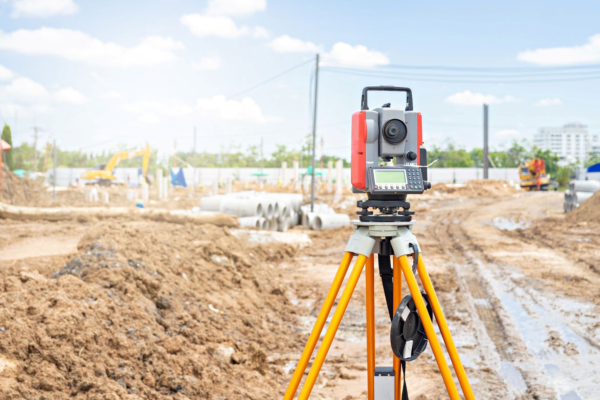 Surveying instrument on a tripod at a muddy construction site under a bright sky