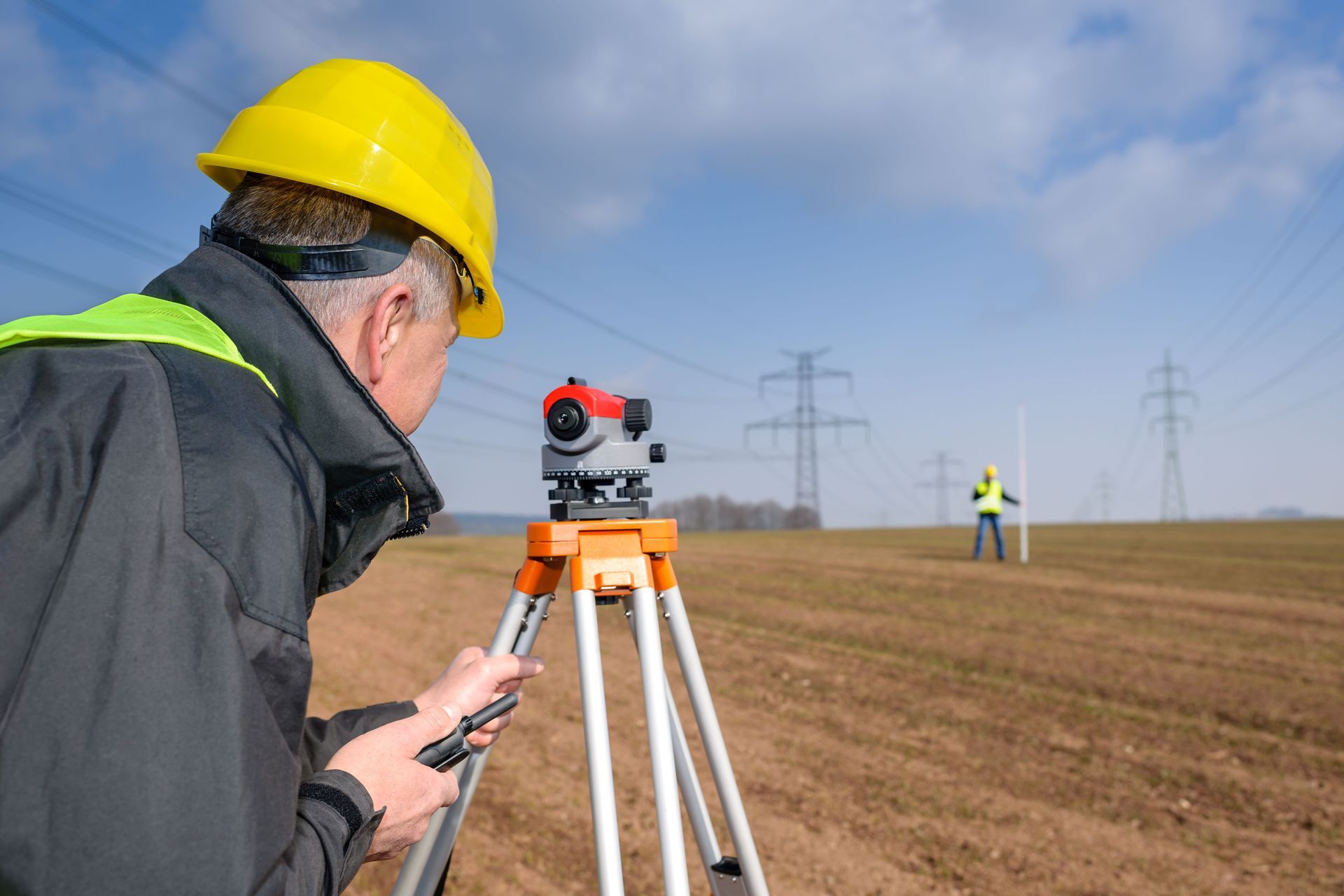 Surveyor in yellow hard hat uses a tripod-mounted instrument in a field with power lines in the background