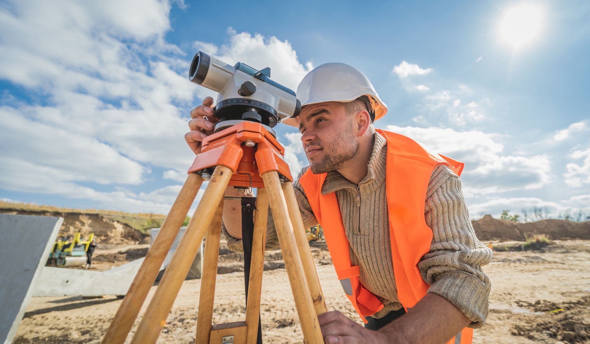 Land surveyor in hard hat and safety vest using a level on a tripod outdoors.