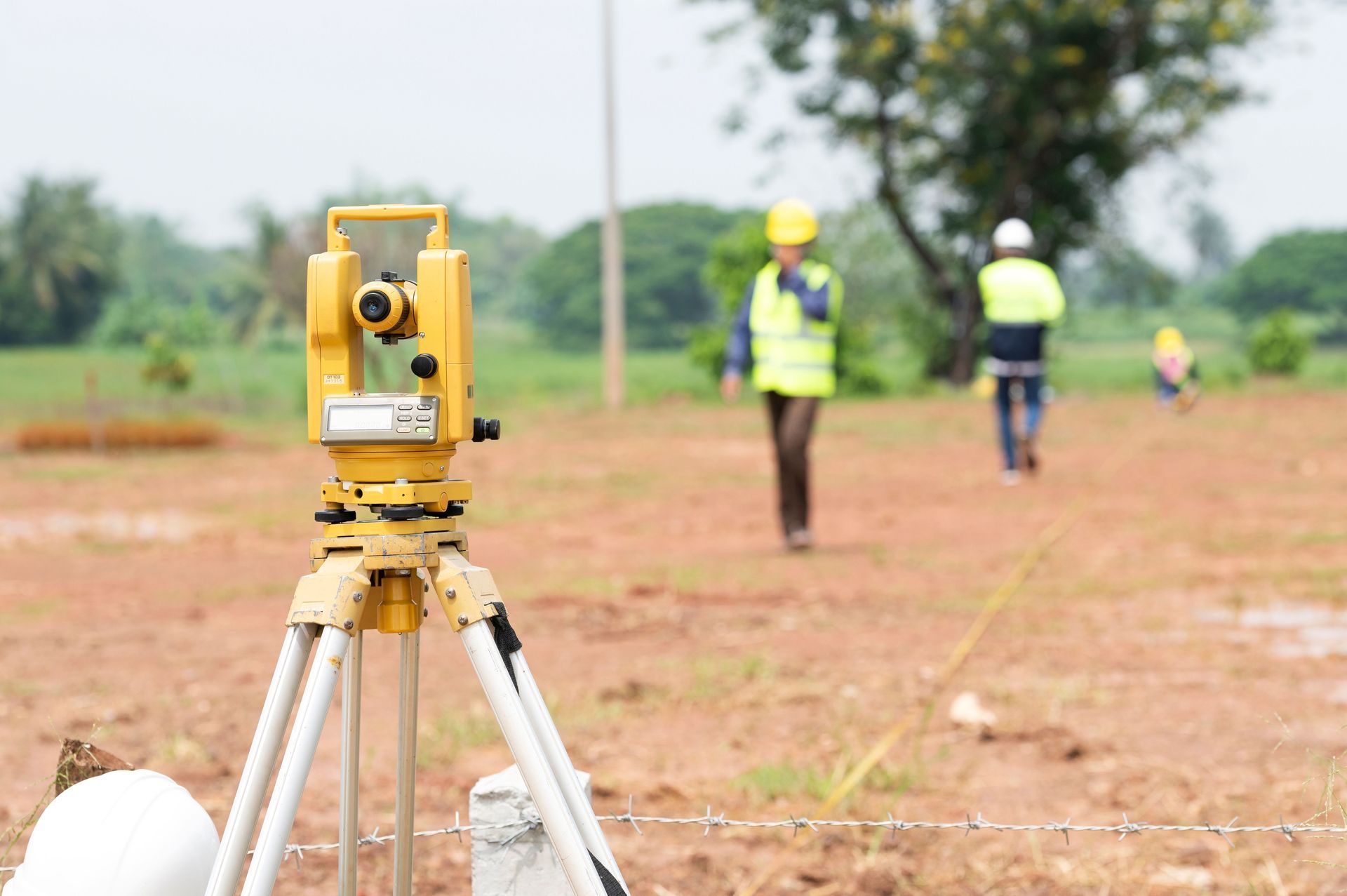 Survey tripod in foreground with workers in yellow vests measuring a dirt field