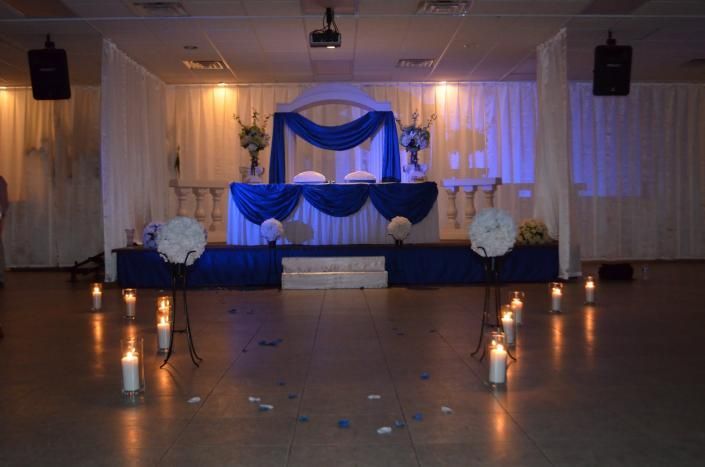 Decorated banquet stage with blue and white drapes, candles, and floral arrangements in a hall