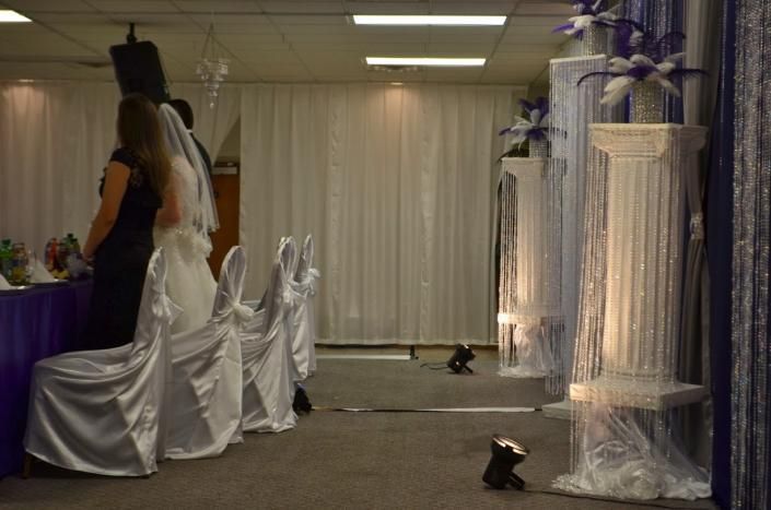 Decorated indoor hall with white draped chairs and curtains, purple accents, and a woman standing at left