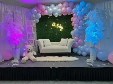 Balloon-decorated stage with white sofa, teddy bear, and pink-blue uplighting against a green backdrop