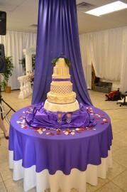 Tiered wedding cake on a purple-draped table in a decorated event hall