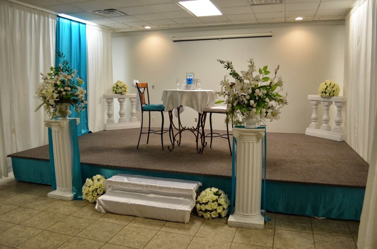 Small decorated stage with a round table, chairs, and floral arrangements in a banquet hall