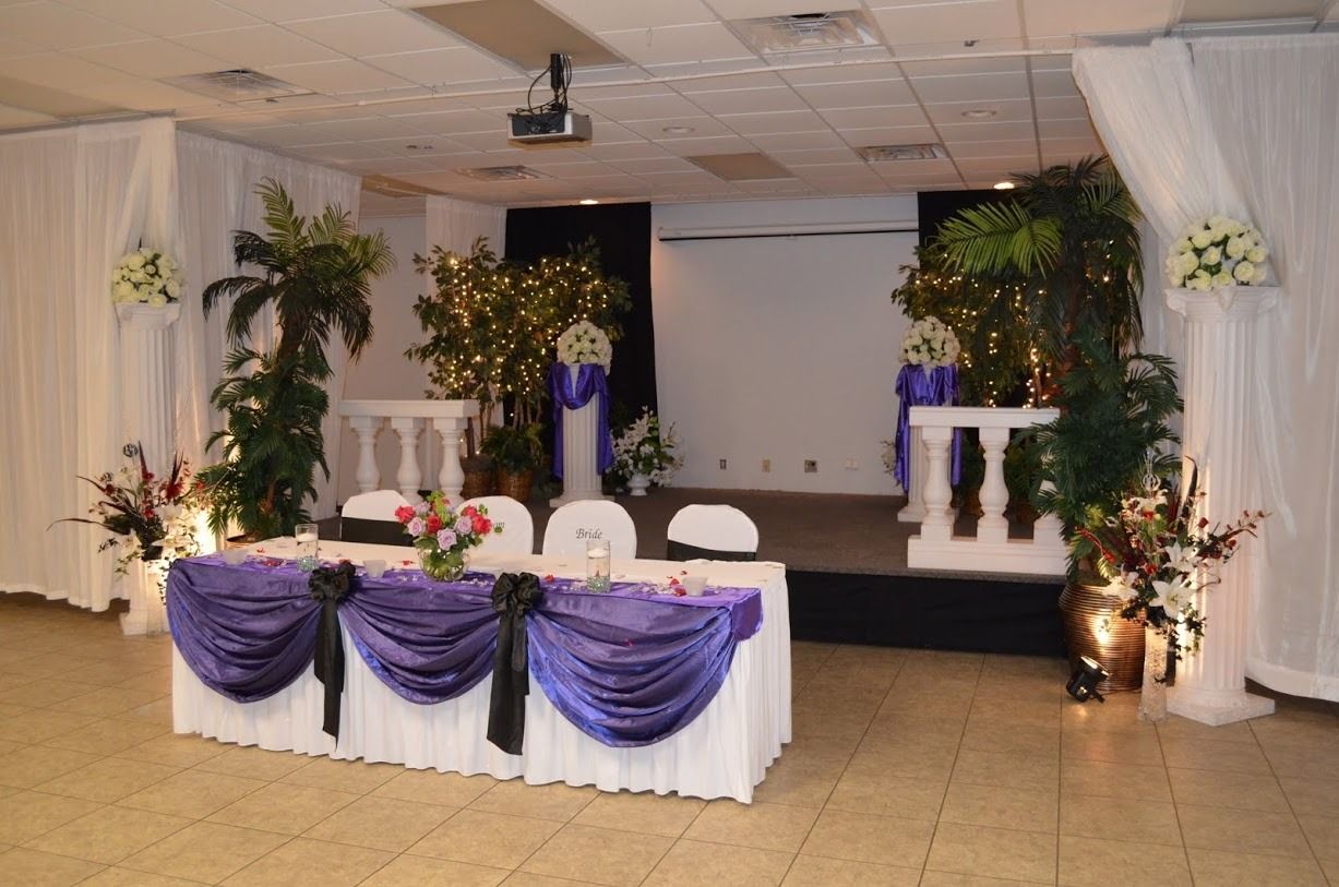 Decorated event stage with purple drapes, floral arrangements, and a long white table with chairs