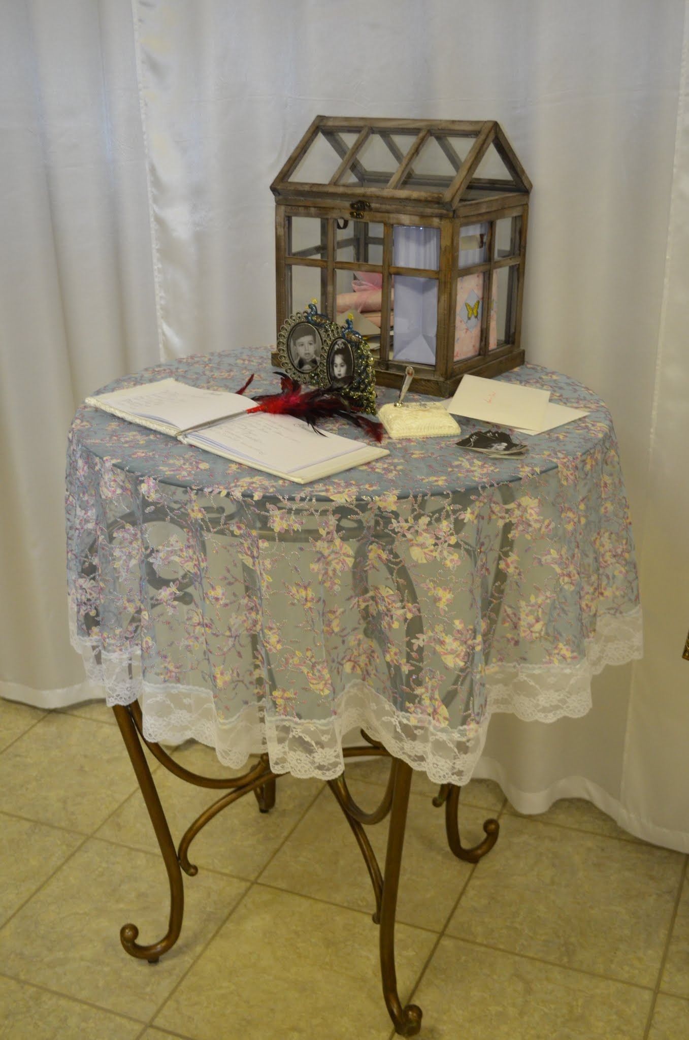 Round table with lace cloth, papers, and a small glass greenhouse on top in a tiled room.