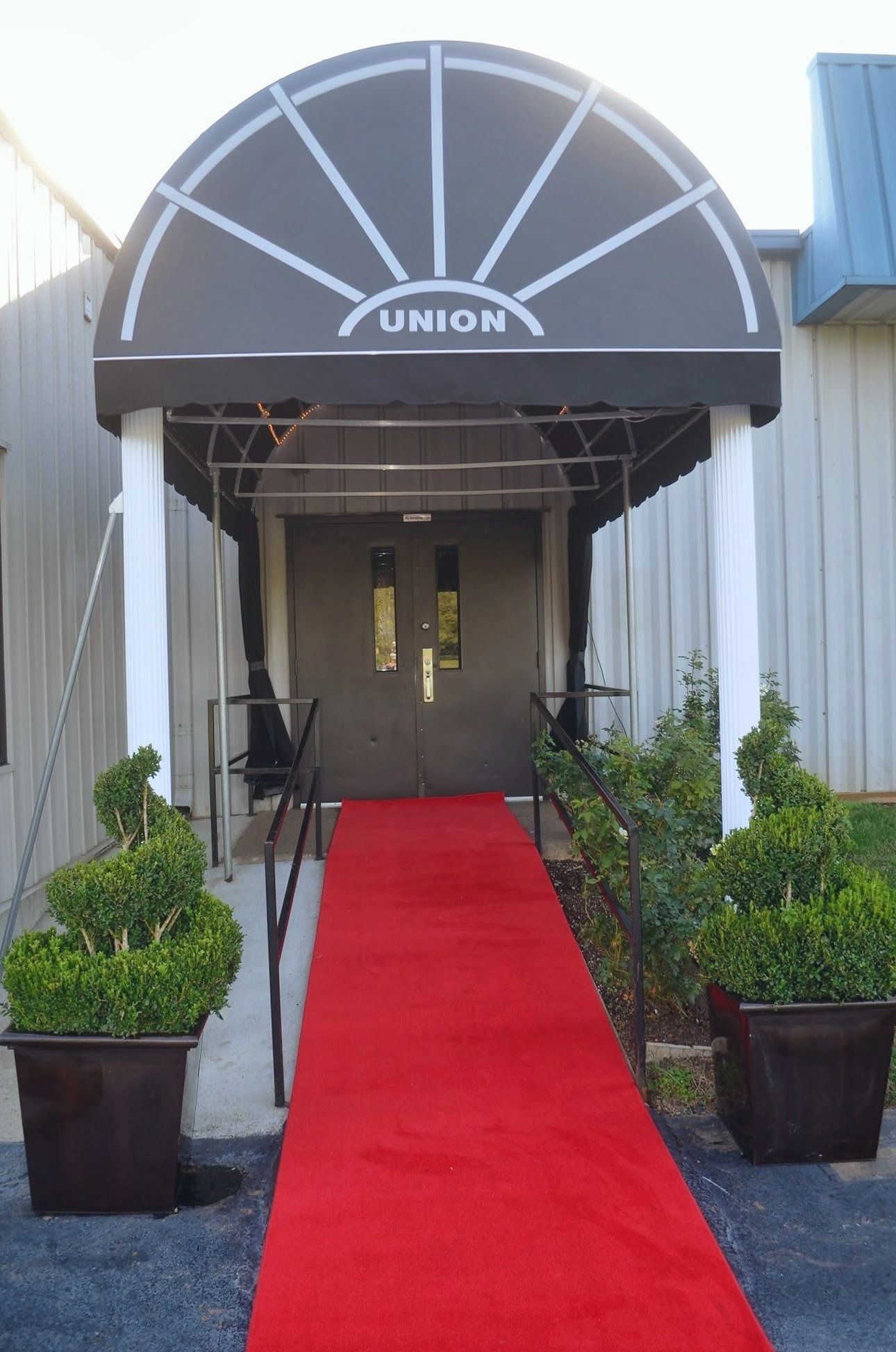 Red carpet leading to a building entrance with a dark awning and potted plants on both sides.