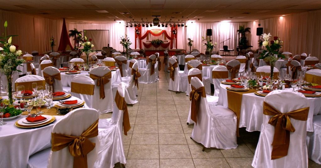 Banquet hall set for a formal event, with white-covered chairs, tables, and a central aisle to a decorated stage