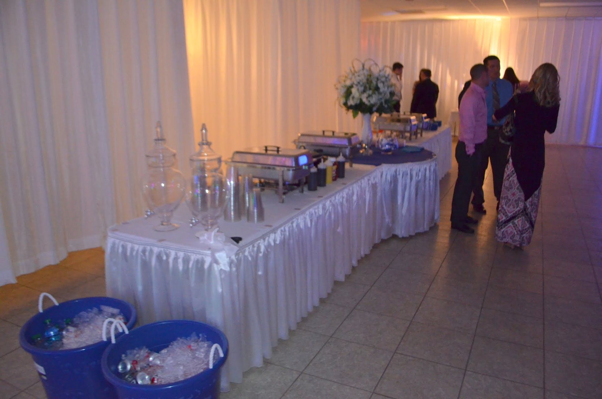 Buffet table with drinks and refreshments at an indoor event, with a few people standing nearby