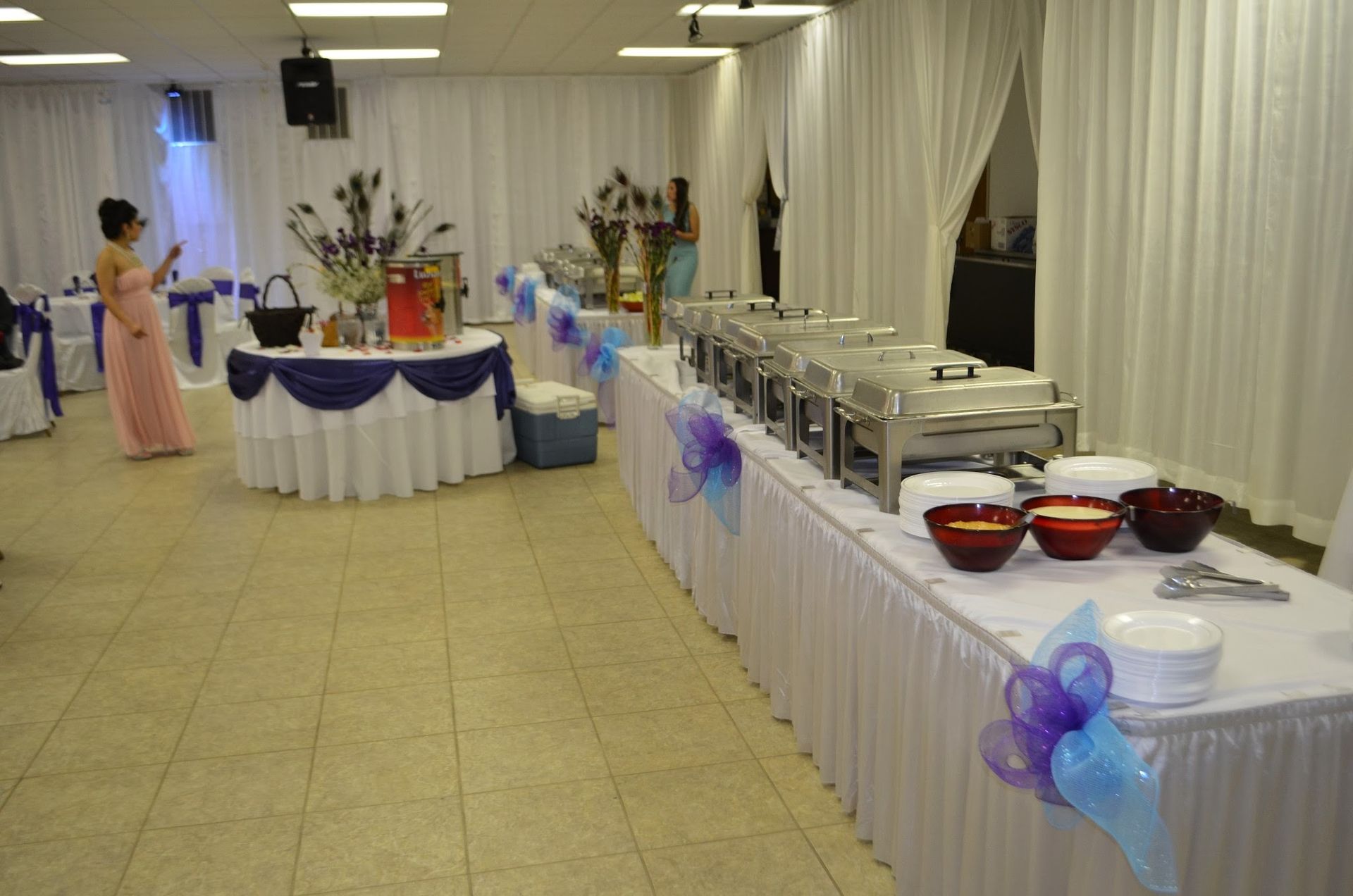 Buffet setup in a decorated banquet hall with white drapes, blue accents, and covered serving trays