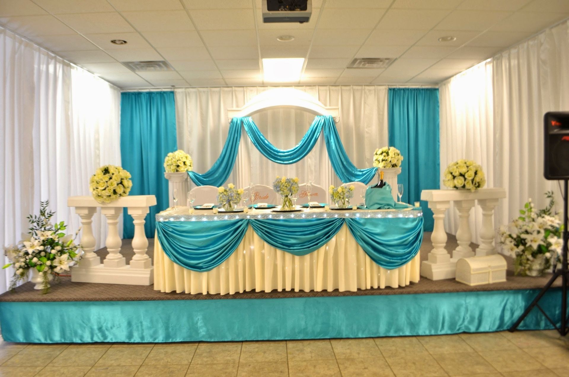 Decorated banquet stage with turquoise and white drapes, floral arrangements, and a long table under bright lights