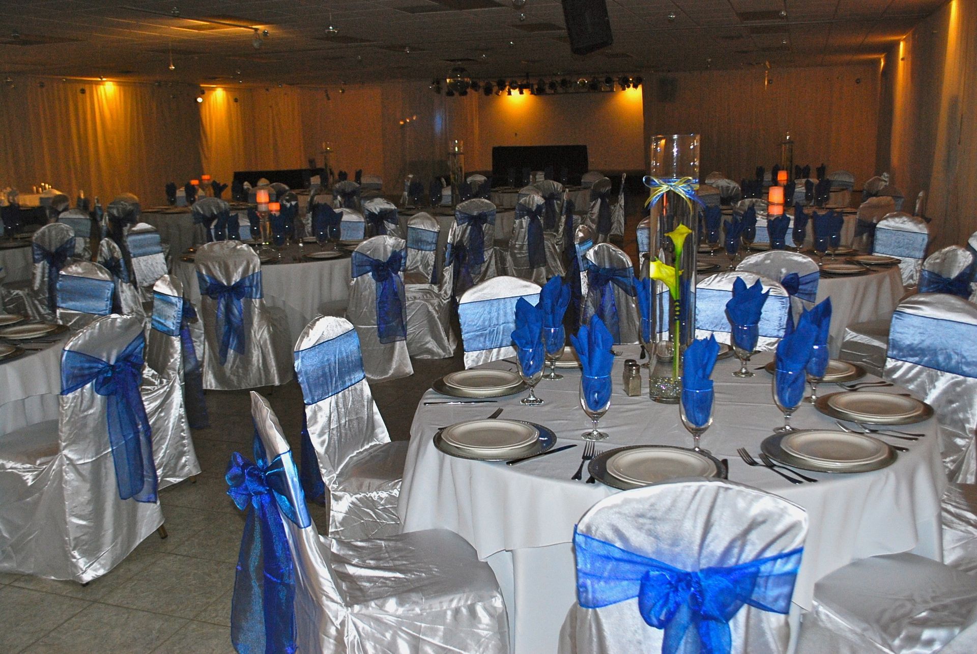 Round banquet hall set for an event, with white chair covers and blue ribbons around round tables and place settings